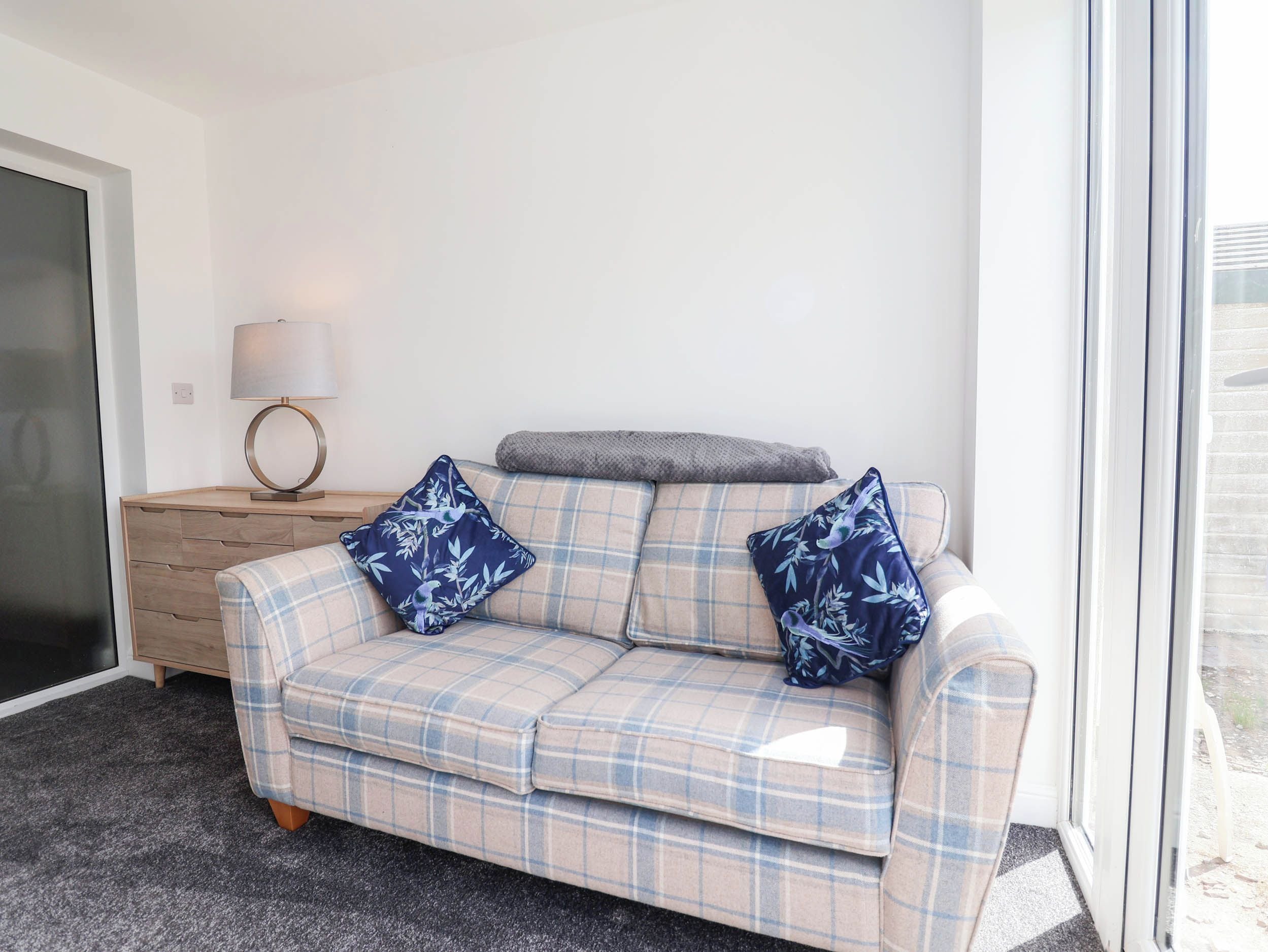 A plaid sofa with floral cushions, a side table with a lamp, and a view through a glass door.