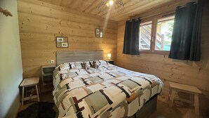 Une chambre dans une cabane en bois, avec un lit, des tables de chevet, un petit tabouret, une fenêtre avec des rideaux et un luminaire au plafond.