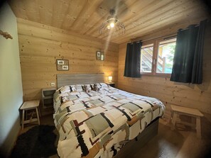 Ein Schlafzimmer in einer Holzhütte mit einem Bett, Nachttischen, einem kleinen Hocker, einem Fenster mit Vorhängen und einer Deckenleuchte.
