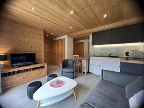 Ein modernes Interieur mit Holzbalkendecke, einem Flachbildfernseher, einer Essecke mit Tisch und Stühlen, einer Couch und einer Küchenzeile.
