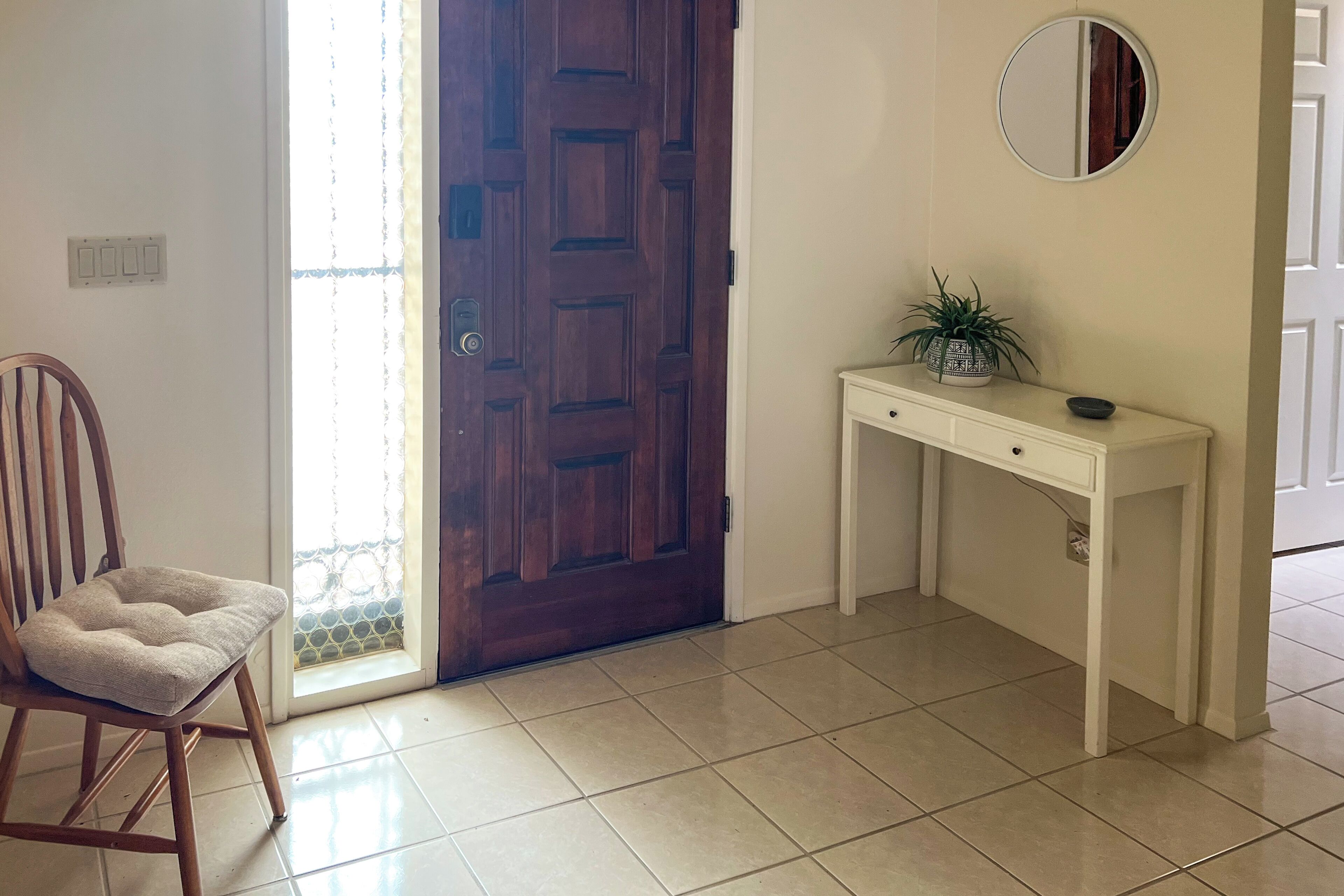 A room with a wooden door, a white console table with a plant, a cushioned chair, and a mirror on the wall.