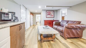 Una cocina con microondas, cafetera y un mueble de madera. Se observa una mesa de comedor con mantel floral y un sofá de cuero marrón. También se ve un armario rojo y una fotografía enmarco.