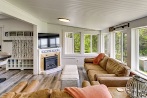 Ein gemütliches Wohnzimmer mit Kamin, Fernseher und großem Fenster mit Blick auf Grünflächen.