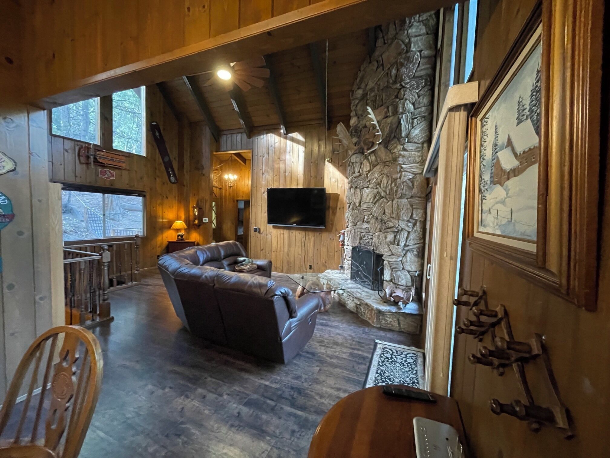 A cozy cabin interior with a stone fireplace, leather sofa, wooden floors, and a mounted TV.