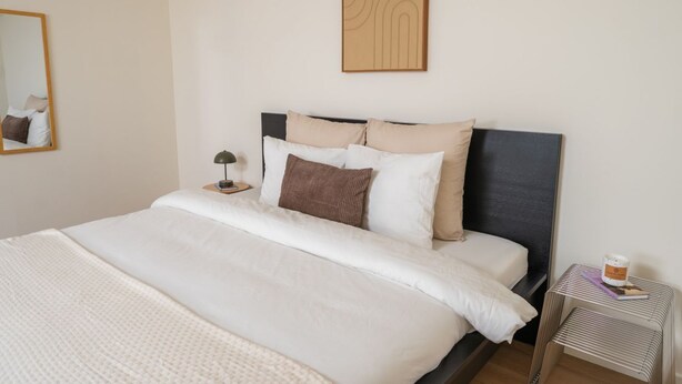 A neatly made bed with a headboard, a bedside table with a lamp, and a small side table with a book and a container. - 15-Min Walk to Ghent Center | Cozy Private Room (Ghent)