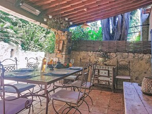 Eine Terrasse im Freien mit Esstisch, umgeben von Stühlen, einer Hängelampe und einer Steinmauer mit Grünpflanzen.