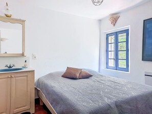 Ein Schlafzimmer mit Bett, Kommode, Spiegel, Fenster mit blauen Fensterläden und einer Wandleuchte.
