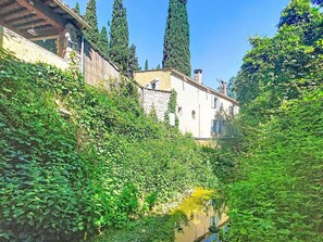 A narrow waterway with dense greenery on both sides, a stone building with a tiled roof, and tall trees in the background. - Le Vieux Moulin by Interhome (Plan de la Tour)