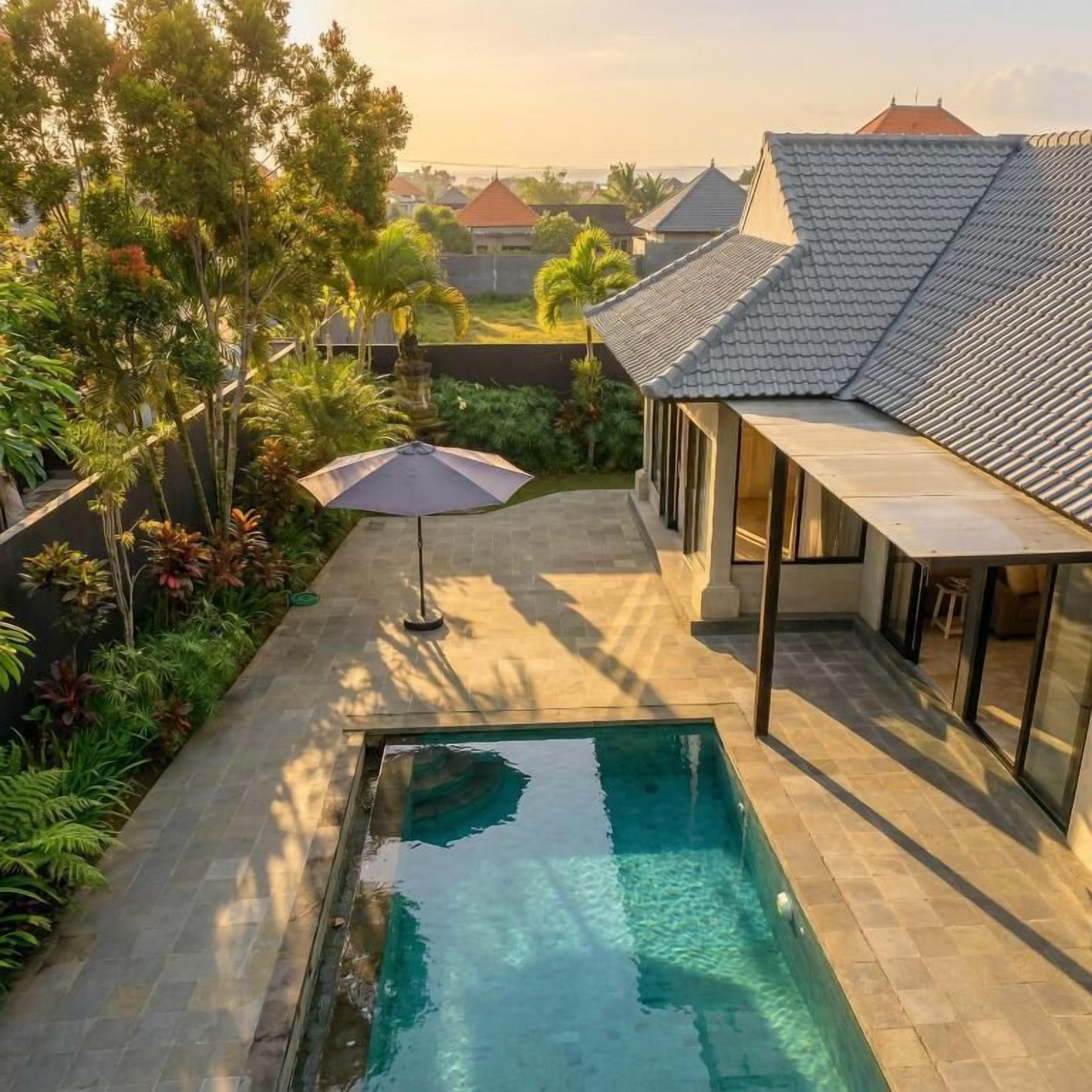 Une piscine avec une terrasse carrelée, entourée d’une végétation luxuriante et une maison avec un toit en tuiles.