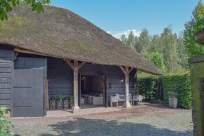 Un bâtiment rustique en bois, doté d’un toit de chaume, d’un espace extérieur couvert aménagé en coin salon et d’une entrée pavée.