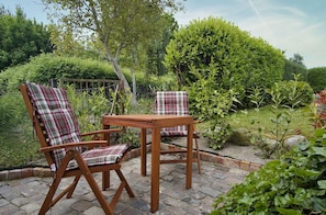 Ein Garten-Terrasse mit zwei Holzstühlen und einem Tisch, umgeben von Grünpflanzen und einer Ziegelumrandung.
