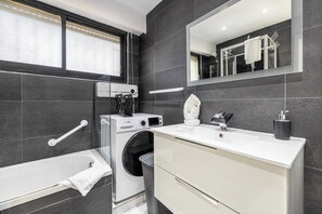 Ein modernes Badezimmer mit integrierter Waschmaschine und Trockner, einer Badewanne, einem großen Spiegel und einem weißen Waschtisch.