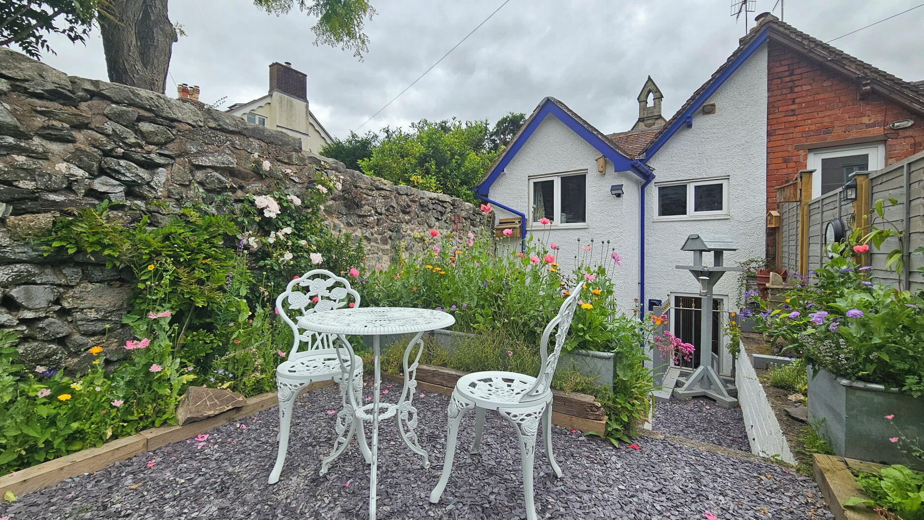 A garden with a stone wall, a white house with a blue roof, and a white metal table and chairs.