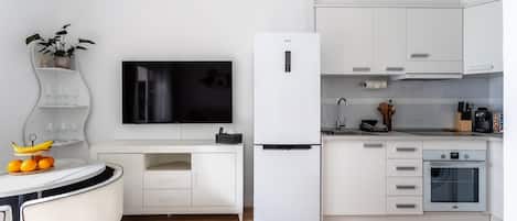 A modern kitchen with white cabinets, a built-in oven, a refrigerator, a flat-screen TV, and a dining area with a table and chairs.