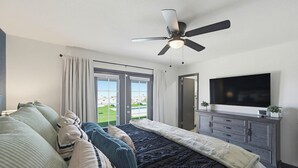 A bedroom with a bed, a ceiling fan, a TV, a dresser, and a view of a green lawn. - Pool, Game Room & Lake View! Modern Havasu Home (Lake Havasu City)