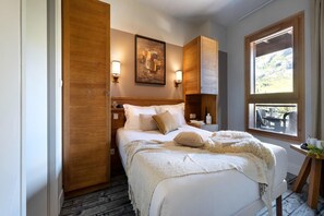 A bedroom with a wooden wardrobe, a bed with white bedding, a painting on the wall, and a window with a view. - Stunning 6-Person Apartment with Balcony (Flaine)