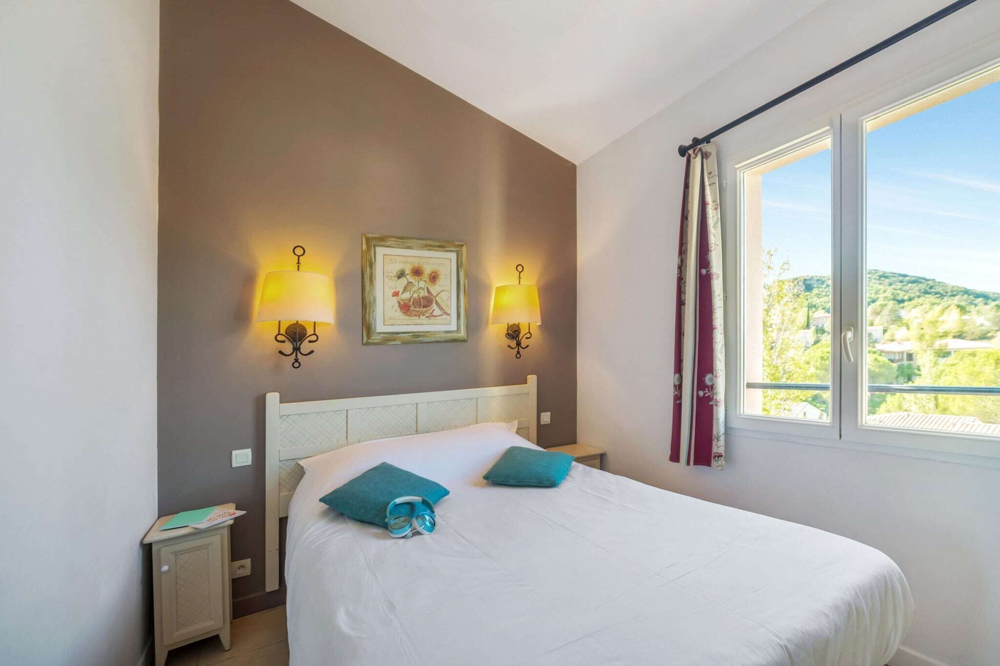 Un dormitorio con una cama, dos lámparas de pared, un cuadro enmarcado, una ventana con vistas y una mesita de noche con un libro.