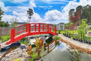 Eine rote Brücke über einen Teich, mit Blumentöpfen und einem Holzzaun im Hintergrund.
