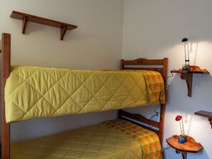 Ein Stockbett mit einer gesteppten, gelben Matratze, ein unteres Bett mit einer karierten Decke, ein Holz-Kopfteil mit einem Symbol und ein Regal mit einer Lampe und einem kleinen Tisch.