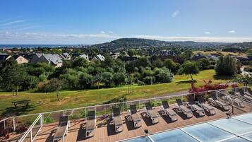 Un espace piscine aménagé sur le toit, avec des chaises longues, une vue sur un quartier résidentiel et une colline au loin.