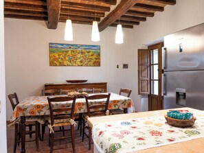 A dining area with wooden chairs, a table with a floral tablecloth, a painting on the wall, and a refrigerator. - "Poggio del Pero" by Interhome (Panicale)