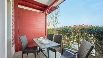 Un balcón con una puerta roja, sillas de mimbre, una mesa con bandeja y vistas a un seto y un edificio.