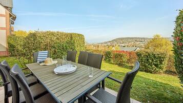 Un patio al aire libre con una mesa dispuesta para cuatro personas, rodeado de sillas de mimbre, con vistas a un jardín ajardinado y una vista distante de un pueblo.