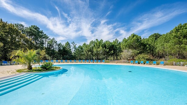 A large, clear blue swimming pool with a wooden deck, surrounded by trees and blue lounge chairs.