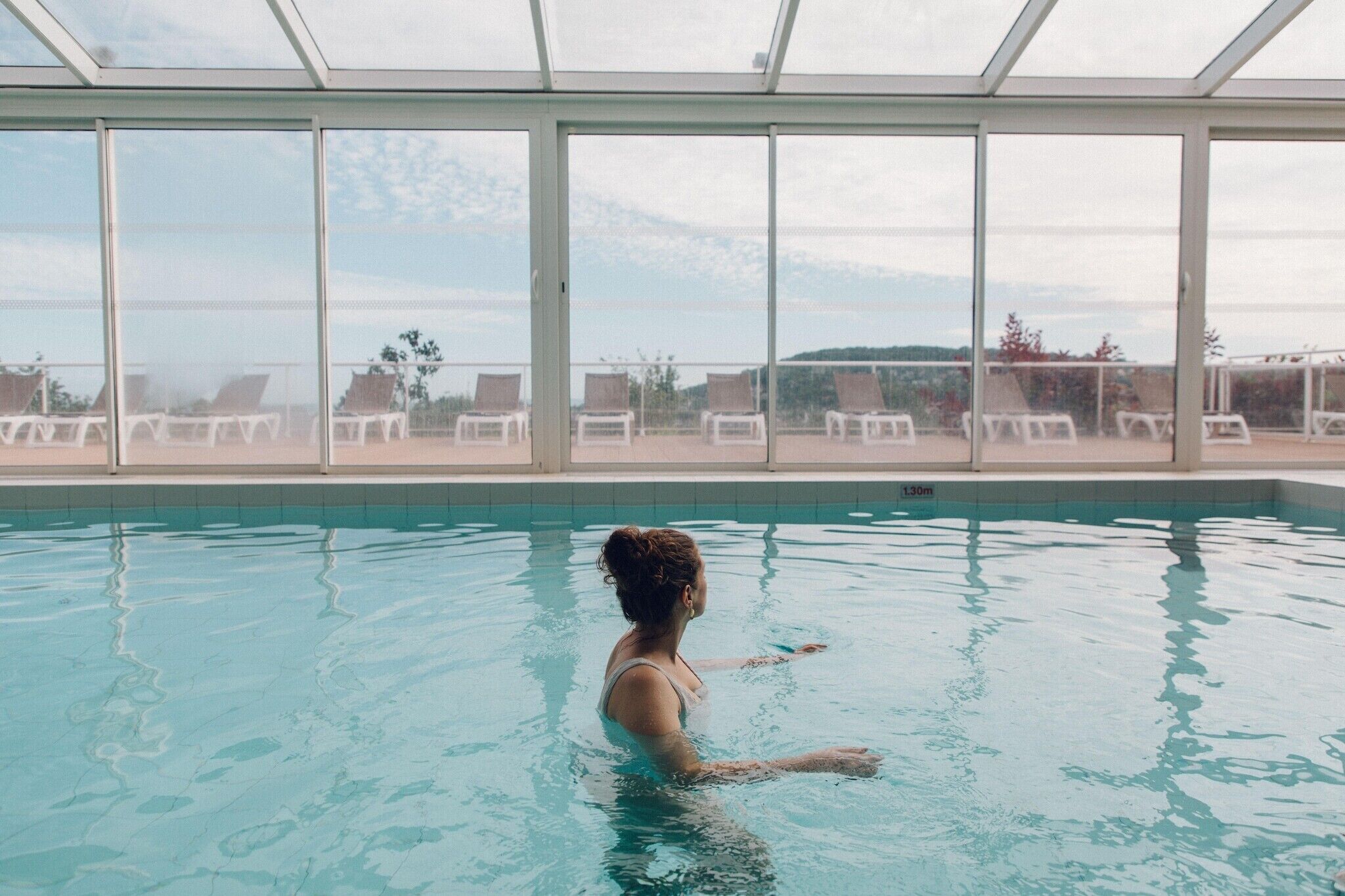 An indoor swimming pool with clear glass walls, a view of the sky, and lounge chairs outside.