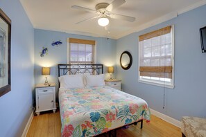 Ein Schlafzimmer mit einem Bett, zwei Nachttischen, einem Deckenventilator, einem Fenster mit Jalousien und einem an der Wand befestigten Spiegel.
