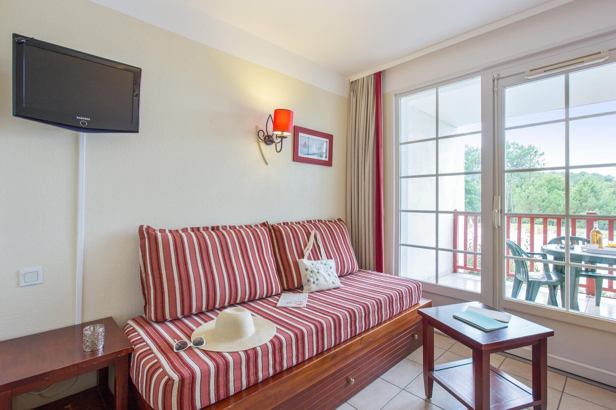Une pièce avec un canapé à rayures, une petite table, un téléviseur fixé au mur et un balcon aménagé avec du mobilier d’extérieur.