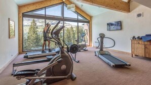 Ett gym med olika typer av träningsutrustning, stora fönster och utsikt över träd.