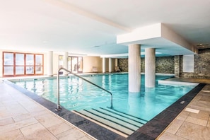 Ein beheiztes Hallenbad mit klarem Wasser, gefliestem Boden und Blick ins Freie durch große Fenster.