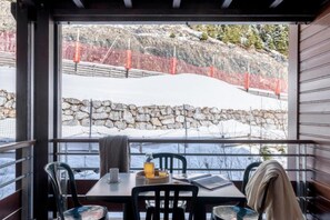 A dining area with a table and chairs, a window with a view of a snowy landscape and a stone wall. - Alps Flat with Heated Pool (Flaine)