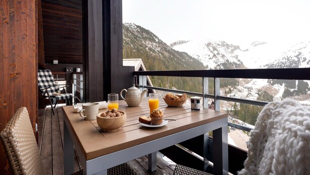 Una mesa de desayuno con alimentos para el desayuno, vistas a montañas nevadas y una barandilla de balcón.