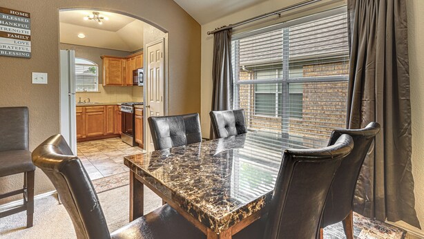 A dining area with a marble table and leather chairs, a kitchen in the background, and a view through a window with blinds. - Fenced Yard & Patio! Family Escape in DFW Suburbs (Fort Worth)