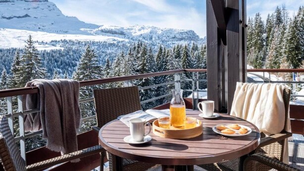 Ein Holztisch mit einer Serviertablett voller Speisen und Getränke, ein Korbstuhl und der Blick auf schneebedeckte Berge.
