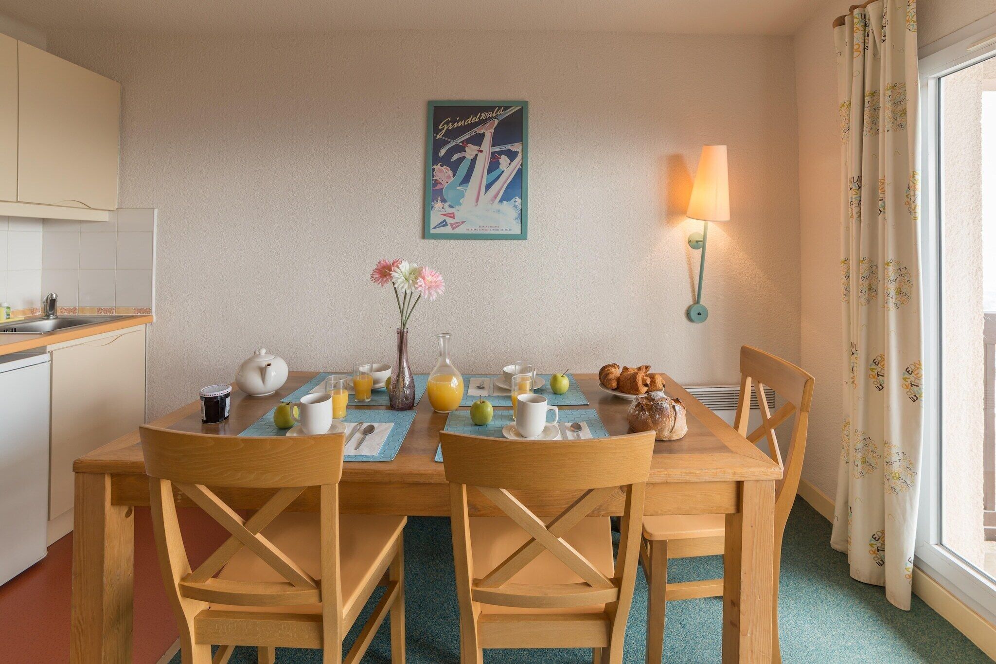 A dining area with a table set for two, a vase with flowers, a lamp, and a poster on the wall.