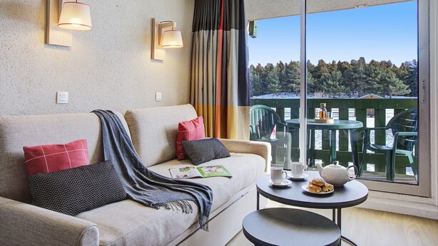 Ein modernes Zimmer mit einer Couch, einem kleinen Tisch mit Teekanne und Keksen sowie einem Balkon mit Stühlen.