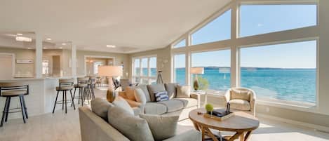 Un vaste espace de vie avec une grande fenêtre donnant sur la mer, un ventilateur de plafond et une cuisine moderne en arrière-plan.