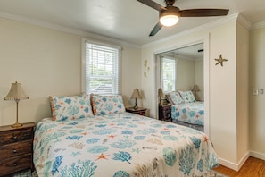 A bedroom with a bed, a ceiling fan, a nightstand, a lamp, a mirror, and a window with blinds.