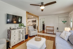 A living room with a television, a ceiling fan, a sofa, a chair, and a kitchen in the background.