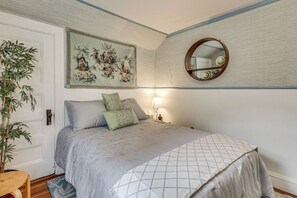 Un dormitorio con una cama, almohadas, una mesita de noche, una lámpara, un espejo y un cuadro decorativo en la pared.