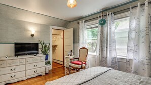 Ein Schlafzimmer mit Bett, Schminktisch, Fernseher, Stuhl und Fenstern mit Vorhängen.