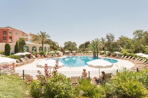 A pool area with sun loungers and umbrellas, surrounded by greenery and buildings.