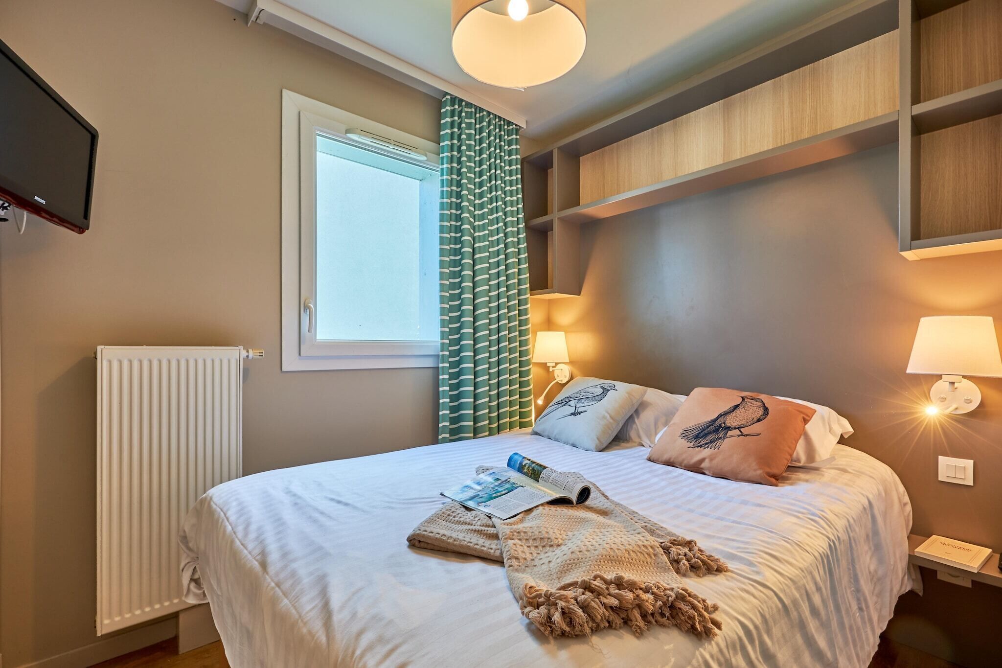 Un lit bien fait, avec un magazine, une couverture tricotée et des oreillers, une lampe sur pied et un téléviseur fixé au mur.