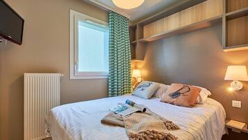 Un lit bien fait, avec un magazine, une couverture tricotée et des oreillers, une lampe sur pied et un téléviseur fixé au mur.