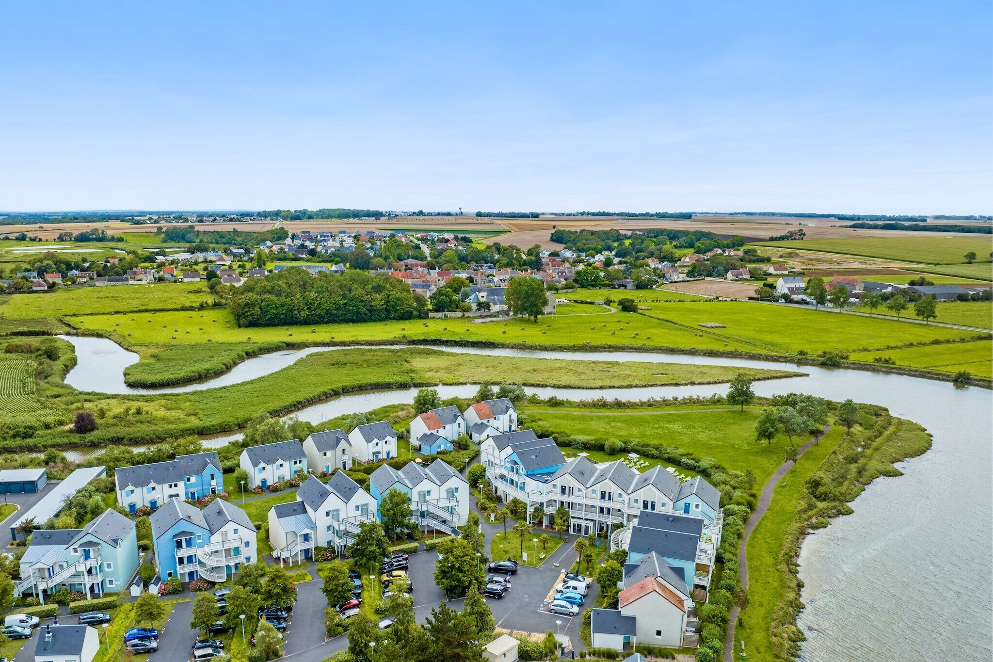 Een woonwijk met huizen aan een rivier, een parkeerplaats en groene velden.