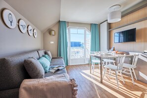 Ein modernes Wohnzimmer mit Sofa, einem Esstisch mit Stühlen, einem flachbildfernseher an der Wand und einem Balkon mit Vorhängen.