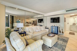 Ein Wohnzimmer mit einer Couch, einem Hocker und einem Couchtisch. Im Hintergrund ist eine Küche mit Schränken und einem Kühlschrank zu sehen.